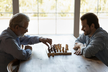 Side view young man with mature father playing chess together, sitting at wooden table at home,...