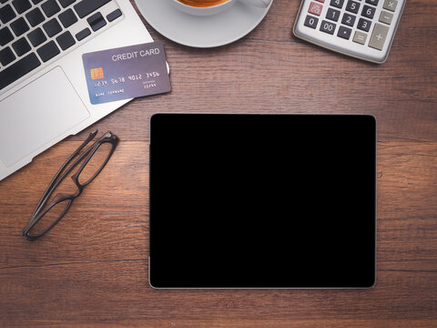 Top view of a tablet with a laptop, credit card, glasses, a coffee cup, and calculator on wooden table in the office. Flat lay, space for text. Working desk table concept