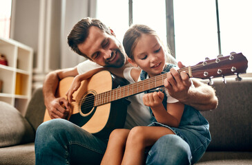 Happy dad teaches his cute daughter to play the guitar while sitting on the sofa in the living room at home. Happy Father's Day. - obrazy, fototapety, plakaty