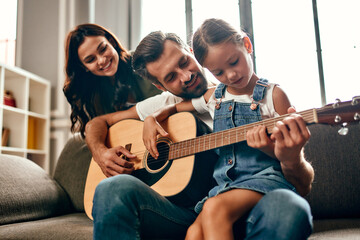 Happy dad and mom teach their cute little daughter to play the guitar while sitting on the sofa in...