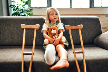 A cute sad little girl with a broken leg in a cast, sits on the couch hugging a teddy bear at home. There are crutches near the sofa for quick rehabilitation.