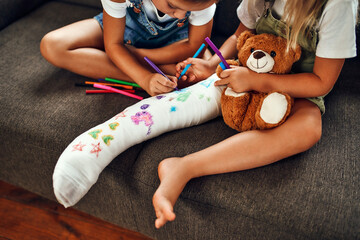 Little girl with a broken leg on the couch. Two sisters draw with felt-tip pens on a plaster bandage. Children have fun and play on the living room couch. - Powered by Adobe