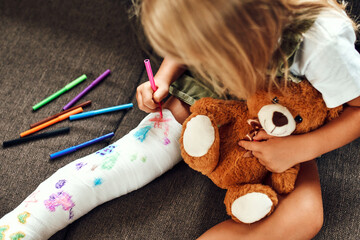Little girl with a broken leg on the couch. The child draws with felt-tip pens on a plaster bandage.