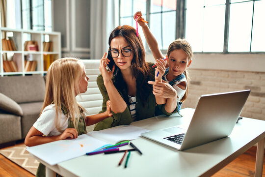 A business woman and a mother are trying to work on a laptop when her little daughters are playing, fooling around and interfering with her. Freelance, work from home.