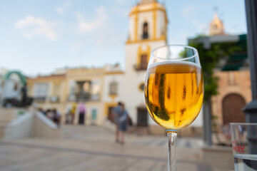 Close-up of a glass full of beer in a famous square outdoors in Ronda, Andalusia, Spain. Blurred facade of a church in the background. - obrazy, fototapety, plakaty