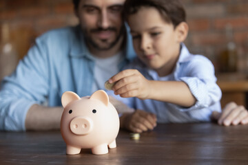 Crop close up of family putting coin into pink piggy bank, caring father teaching little son kid to save money for future, sitting at wooden table at home, insurance and investment concept