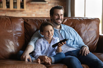 Happy father with little son watching television show series or movie, relaxing sitting on cozy couch at home together, smiling 8s boy kid holding tv remote, switching channel, enjoying leisure time