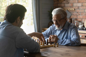 Smiling mature man in glasses with grownup son playing chess together, sitting at wooden table,...