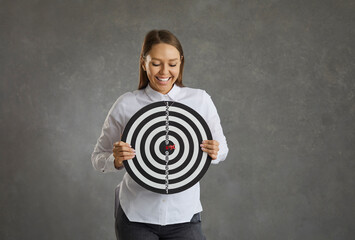 Happy smiling young business lady standing on gray studio background holding shooting target with...
