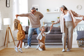 Active senior grandparents dancing with two happy kids grandchildren in living room - obrazy, fototapety, plakaty