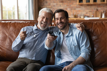 Laughing young man with mature father watching tv together, hugging, relaxing on couch at home,...