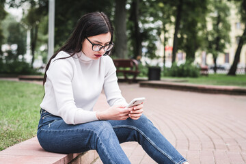 Beautiful stylish girl in jeans and a white sweater outdoors in the park with a phone