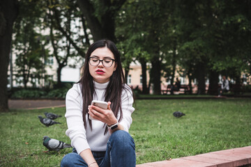 Beautiful stylish girl in jeans and a white sweater outdoors in the park with a phone