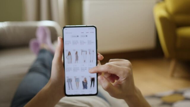 Woman Looks at Goods in Online Store Clothing. Buy Fashion Clothes Directly on Smartphone. Woman at Home Lying on Couch in Living Room Using Smartphone Buys in Internet Shop.