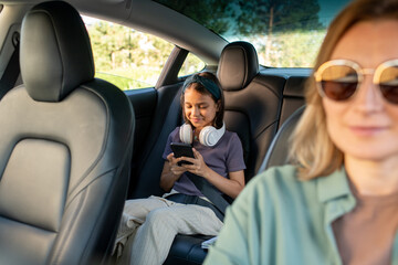Happy schoolgirl with headphones scrolling in smartphone while sitting on backseat of car