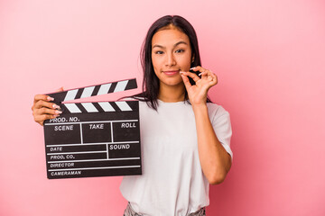 Young latin woman holding clapperboard isolated on pink background  with fingers on lips keeping a...