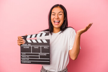 Young latin woman holding clapperboard isolated on pink background  receiving a pleasant surprise,...