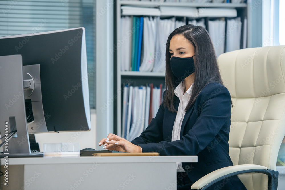 Aufkleber young serious secretary in protective mask looking at computer screen during network in office