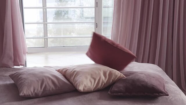 Four decorative pillows falling on bed. Someone throws cushions on sofa. Cozy interior with large window and curtains in maroon, pale violet and pink color tones.