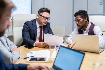 Young serious businessman lookng at paper while mature colleague or boss explaining data