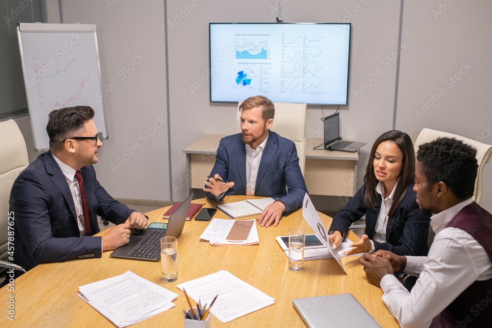 Poster Two businessmen consulting about speech points while two intercultural colleagues discussing papers by table in boardroom