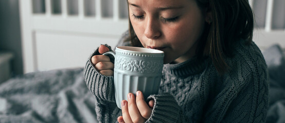 Young girl drinking hot chocolate on cozy bed in bedroom. Concept of autumn, winter weekend. Banner