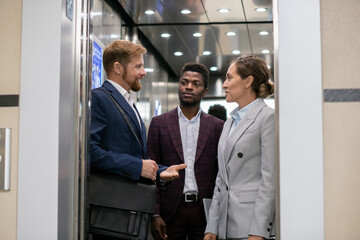 Young African businessman looking at his interacting colleagues in elevator