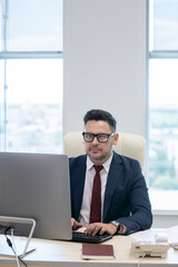 Busy man in formalwear typing on computer keyboard while sitting by desk in front of monitor