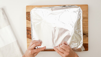 Woman cover baking pan with aluminum foil, close up view from above, white kitchen table background
