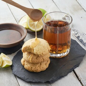 Vaso de t&eacute; con galletas artesanales de mantequilla con miel natural de abeja y rodajas de lim&oacute;n sobre un plato de piedra laja y fondo de madera blanca.