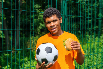 Side view portrait of young African-American man holding soccer ball while standing against fence in sports court outdoors, copy space