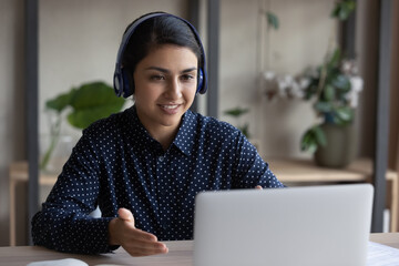 Smiling Indian woman in wireless headphones chatting online by video call, looking at laptop screen, friendly businesswoman engaged in virtual event, teacher mentor coach leading webinar, explaining