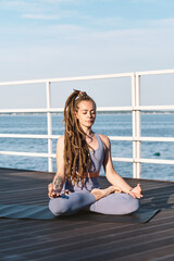 Contemporary sportswoman sitting in pose of lotus on pier against blue sky and water