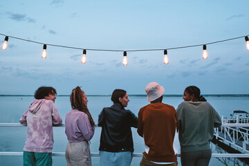 Group of young friends looking at African girl during conversation by waterside on summer evening
