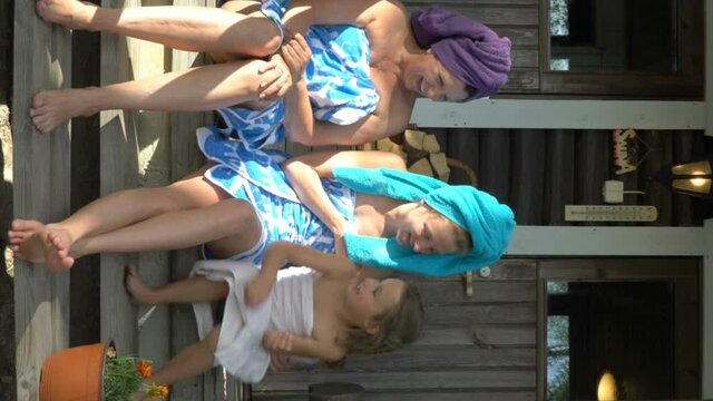 Young woman, her mature mom and her little daughter having a fun enjoying the silence and fresh air on the porch of a traditional Finnish sauna wearing towels. A vertical video.