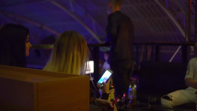 A young woman in a nightclub takes a selfie on her phone, sitting at a table. Against the background of her group of people, guys and girls are drinking and having fun. Restaurant or bar.