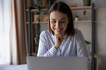 Fototapeta premium Smiling businesswoman or student using laptop, looking at screen, working online, happy young female watching webinar or video, reading good news in message, enjoying leisure time with computer