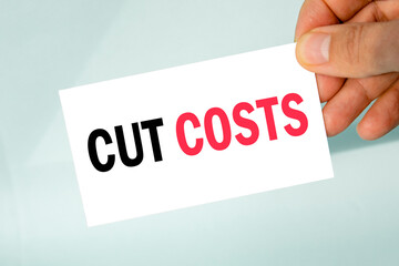 businessman's hand holding paper business card with text cut costs, closeup light gray background