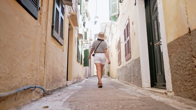 Back view of tourist woman walking alone, exploring old european city narrow streets on Greece island. Lady sightseeing local architecture. She is having good time wandering around town in summer