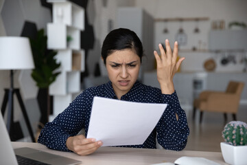 Unhappy annoyed Indian woman reading bad news in letter, sitting at home office desk, upset...