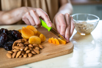 female hands preparing a healthy breakfast: cut dried fruits and nuts with a green knife: dried apricots, almonds and dates on a wooden cutting board into a glass bowl