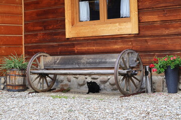 Czarny kot pod ozdobną ławką, przy drewnianym górskim domu. Black cat under the bench at the mountain cottage - obrazy, fototapety, plakaty