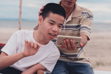 Happy disabled teenager boy smiling face with parent using mobile phone on outdoors activity at the beach, Vacation of people with disabilities and family in nature outside , Mental health concept. - Powered by Adobe
