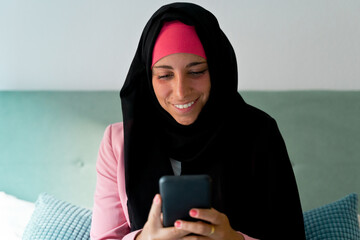 Cheerful muslim woman wearing a hijab using smartphone indoors. Horizontal side view of arabic...