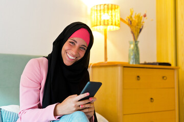 Cheerful muslim woman wearing a hijab using smartphone indoors. Horizontal side view of arabic woman using technology in bed at home. Technology and muslim women lifestyle