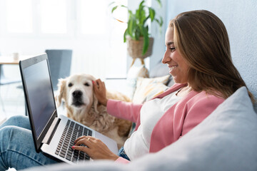 Cheerful blonde woman working on computer with pet on the couch at home. Horizontal side view of caucasian woman playing with her dog indoors. Technology, animals and people lifestyle.
