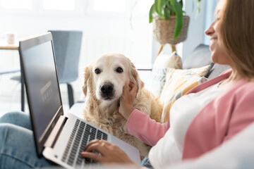 Cheerful blonde woman working on computer with pet on the couch at home. Horizontal side view of caucasian woman playing with her dog indoors. Technology, animals and people lifestyle.