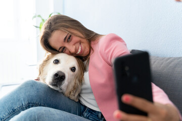 Cheerful blonde woman taking a photo with pet on the couch at home. Horizontal side view of caucasian woman playing with her dog indoors. Technology, animals and people lifestyle.