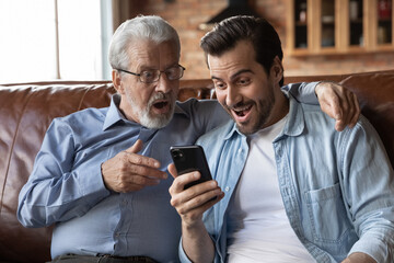 Overjoyed surprised two generations of men looking at phone screen, excited senior father and...