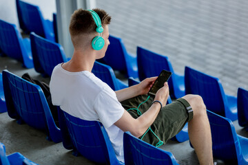 Young teen boy with headphones sitting in blue sit on stadium tribune